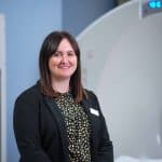 Corporately-dressed person standing in front of a PET-CT scanner in a clinical setting.