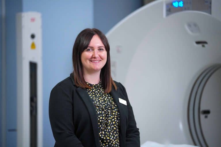Corporately-dressed person standing in front of a PET-CT scanner in a clinical setting.