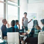 Group of professionals in a modern office engaged in a leadership training session, with a facilitator presenting strategies on a whiteboard.