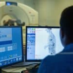 Medical professional monitoring PET-CT scan results on computer screens in a control room, with the scanner visible in the background.