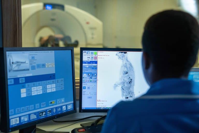 Medical professional monitoring PET-CT scan results on computer screens in a control room, with the scanner visible in the background.