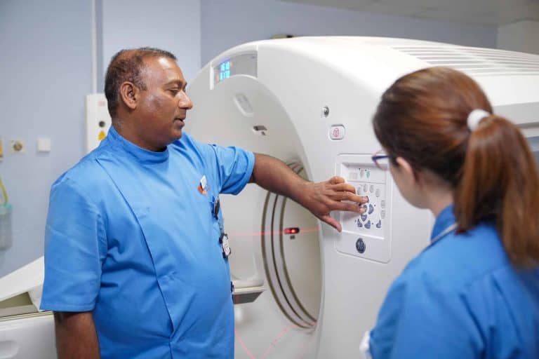 Two healthcare professionals in blue scrubs standing beside a PET-CT scanner, with one pointing at the machine’s control panel during a training or operational procedure.