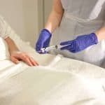 Healthcare professional wearing gloves preparing an IV injection for a patient lying in a hospital bed.