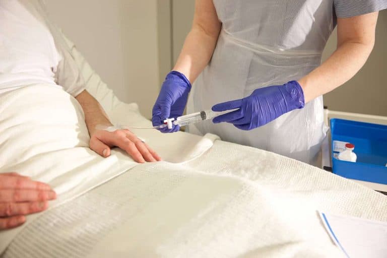 Healthcare professional wearing gloves preparing an IV injection for a patient lying in a hospital bed.