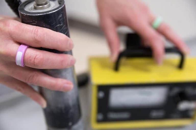 Close-up of hands operating a radiation detection device, with one hand holding a cylindrical probe and the other adjusting a yellow control unit featuring a digital display
