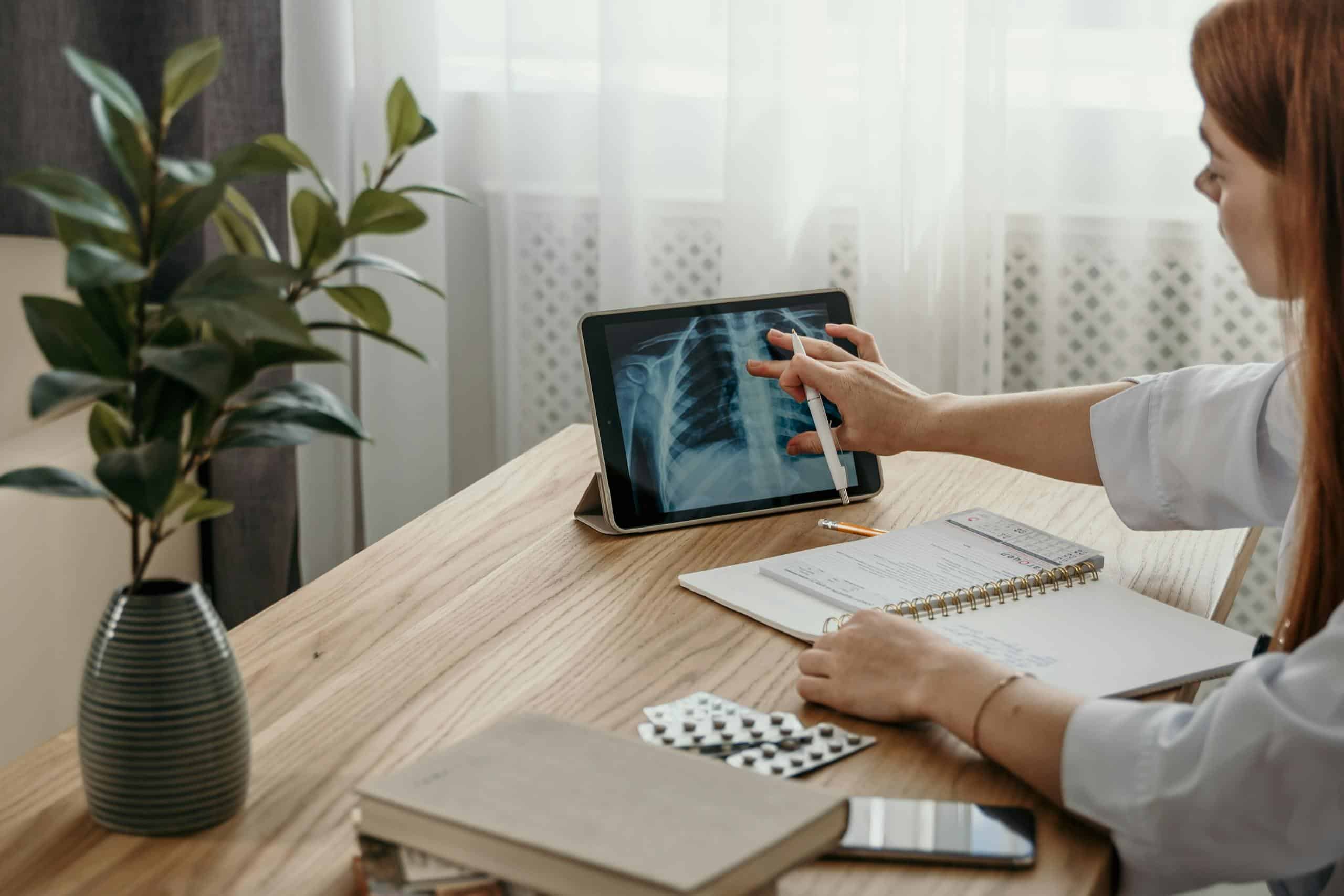 Healthcare professional examines X-ray image on tablet at desk with medical notes.