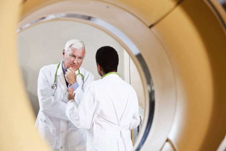 Two healthcare professionals wearing white lab coats and stethoscopes standing near a PET-CT scanner, viewed through the circular opening of the machine during a discussion.