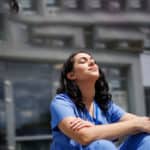Person wearing blue clinical scrubs seated indoors with arms resting on knees, in a modern healthcare environment