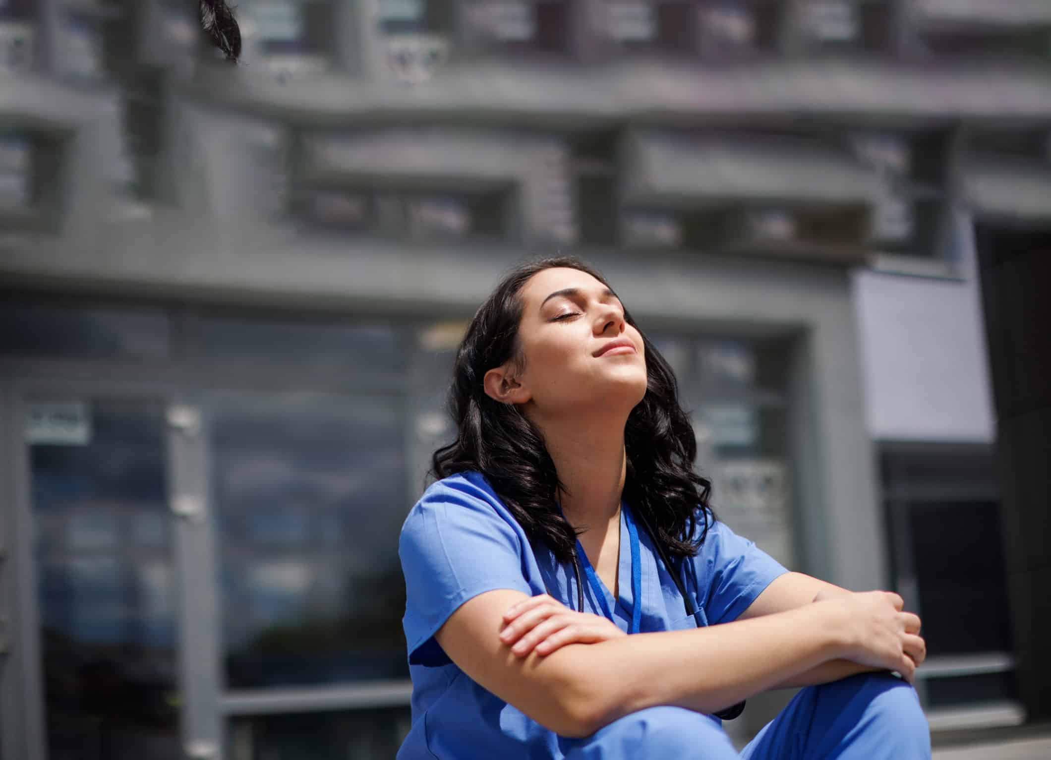 Person wearing blue clinical scrubs seated indoors with arms resting on knees, in a modern healthcare environment
